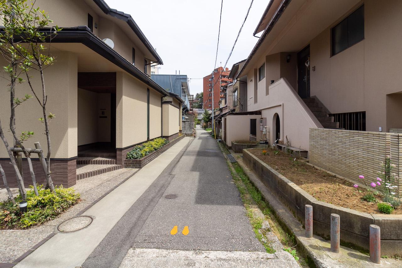 金沢市橋場町の賃貸物件/車１台が通れる幅の前面道路。左側に見える青色の建物が本物件。近隣には比較的新しいお家もちらほら